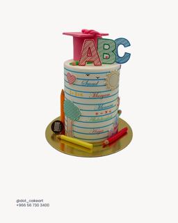 كيكة بشكل دفتر مسطر بحروف ABC وقبعة وردية وأقلام رصاص ومكتوب عليها أسماء من دوت كيك آرت