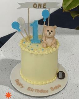 Pale yellow first-birthday cake with teddy bear and balloons, by DOT Cake Art