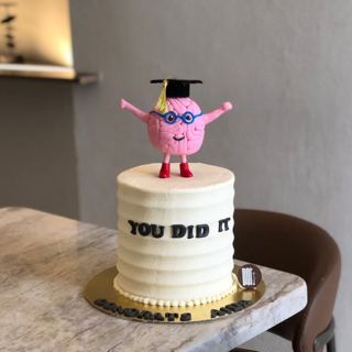 White cake with a brain character in a grad cap, by DOT Cake Art
