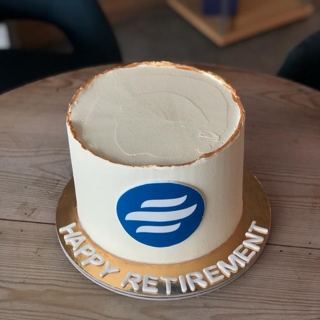 كيكة بيضاء بحافة ذهبية وشعار أزرق وعبارة Happy Retirement من دوت كيك آرت