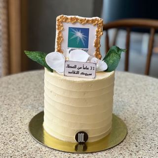 White cake with Aramco logo in gold frame, by DOT Cake Art