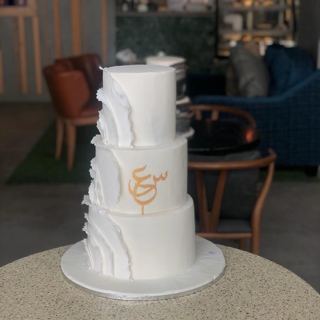 كيكة ثلاث طبقات بيضاء بكشكشة جانبية وزخرفة خط عربي ذهبي من دوت كيك آرت